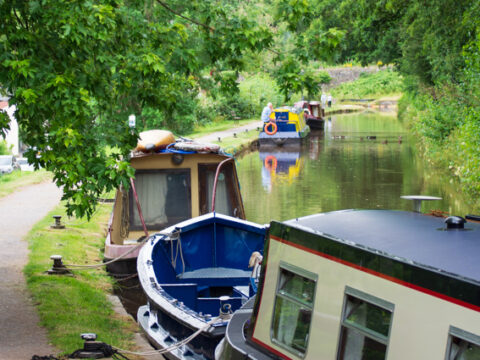 A summer view of people walking along the towpath alongside moored boats on the canal at Talybont. The scene has lots of trees overhanging the canal.