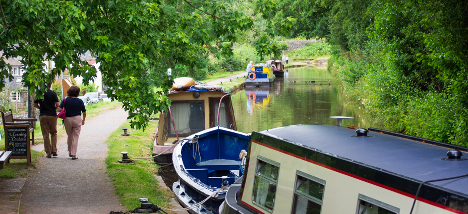 A summer view of people walking along the towpath alongside moored boats on the canal at Talybont. The scene has lots of trees overhanging the canal.
